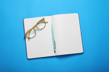 Wall Mural - Open notebook, pen and glasses on light blue background, top view