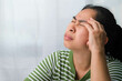 © Pornpimon - Young woman having headache against white curtain background in room. An adult woman touching head because of headache or migraine. Hands on head.
