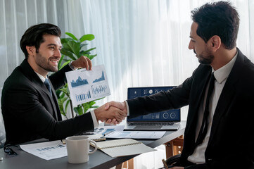 Closeup professional businessman shaking hands over desk in modern office after successfully analyzing pile of dashboard data paper as teamwork and integrity handshake in workplace concept. fervent