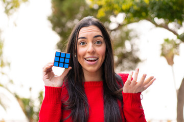 Poster - Young woman holding a three dimensional puzzle cube at outdoors with shocked facial expression