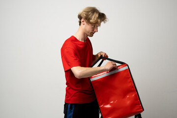 Wall Mural - Young courier opening thermal bag on white background. Food delivery service. Delivery guy in a red t-shirt uniform work as courier and holds red thermal food backpack. Service concept.