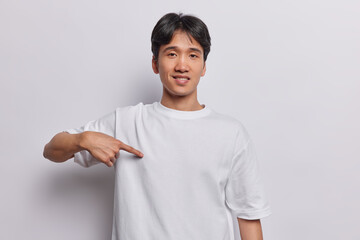 Wall Mural - Clothing and design concept. Cheerful dark haired Asian man points at blank space of t shirt suggests to place your promo designing content or advertisement here isolated over white background