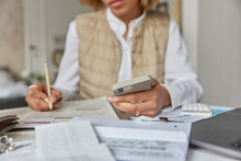 Cropped Shot Of Unrecognizable Woman Holds Mobile Phone Checks Documents Utility Bills Writes Down Information In Notepad Poses At Table Indoors Surrounded By Paper Invoices Counts Home Expenses