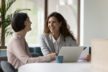 Wall Mural - Two happy businesswomen, office employees work in office, laughing, joking during workflow, share creative ideas, solution feel satisfied with corporation, engaged in teamwork. Negotiation, business