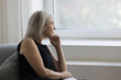 © fizkes - Thoughtful depressed mature woman sitting on couch at home, looking at window away in deep thoughts, suffering from depression, apathy, emotional crisis, thinking over problem, loss