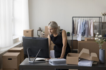 Serious mature internet store owner using desktop computer in small storage place, writing notes, processing order, sending purchase from webstore office, preparing product for packing, packaging