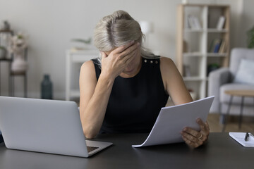 Depressed sad mature entrepreneur, business woman reading paper document at home, sitting at work table with laptop, learning bad shocking news, covering face, getting problems, stress