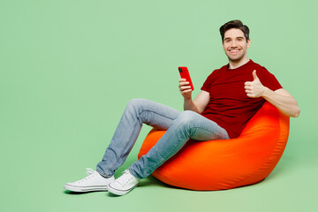Wall Mural - Full body smiling fun young man he wears red t-shirt casual clothes sit in bag chair hold in hand use mobile cell phone show thumb up isolated on plain pastel light green background studio portrait.