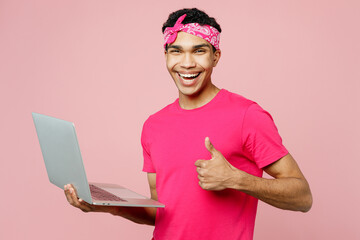 Wall Mural - Side view young IT man of African American ethnicity he wears headscarf t-shirt casual clothes hold use work on laptop pc computer show thumb up isolated on pastel plain light pink color background.