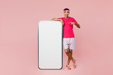 Full body young man wear headscarf t-shirt casual clothes point index finger on big huge blank screen mobile cell phone smartphone with copy space area isolated on pastel plain light pink background.