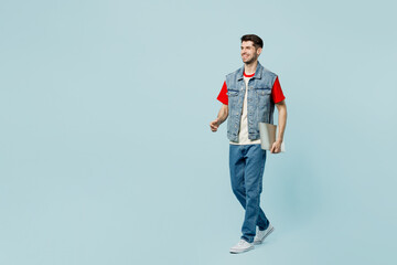 Full body smiling happy young IT man he wears denim vest red t-shirt casual clothes hold closed laptop pc computer walk go isolated on plain pastel light blue cyan background studio. Lifestyle concept