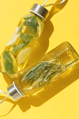Wall Mural - Top view closeup two glass bottles, water drink detox with lemon and mint leaves, and rosemary, at sunlight on yellow color background. Infused water, wellness trend. Refreshing summer drink