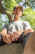 © Valeria Venezia - Low angle view of a young man, serious, sitting on a tree while looking at camera