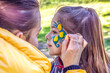 © Nataliia - An artist's hand draws a blue-yellow flower on the face of a little girl. A child with a funny face painting. Children's holiday.