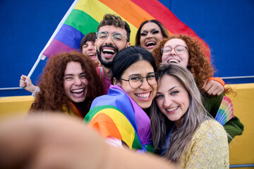 Selfie of a LGBT group of young people celebrating gay pride day holding rainbow flag together. Homosexual community smiling and taking cheerful self portrait. Lesbian couple and friends generation z