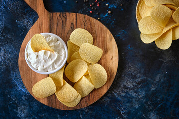 Top view potato chips, sauce in a white bowl on a wooden cutting board.