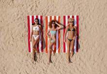 Three Beautiful Young Women Lying On The Beach Sunbathing In Swimming Suites, Relaxing And Enjoying.