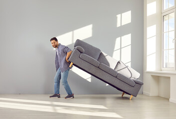 Man moving the sofa at home. Full shot of a happy young man moving his gray couch while moving the furniture and furnishing his new modern house or apartment