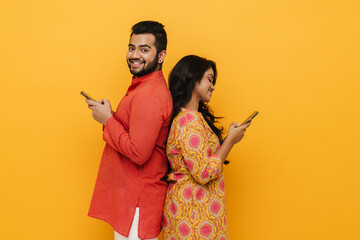 Indian man and woman using mobile phones while standing back to back isolated over yellow background