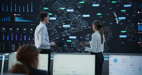 Wall Mural - Male And Female Logistics Specialists Discussing New Routs Of Delivery In Front Of Big Digital Screen In Monitoring Office. Diverse Employees Working Behind Desktop Computers For Startup Service.