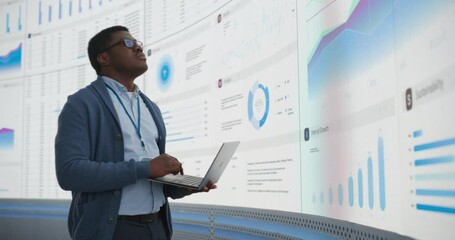 Fototapete - Confident Black Businessman Holding Laptop And Reviewing Financial Reports On Big Digital Screen In Monitoring Office. Male Entrepreneur Analyzing Growth And Profit Margins Of Tech Start Up Company.