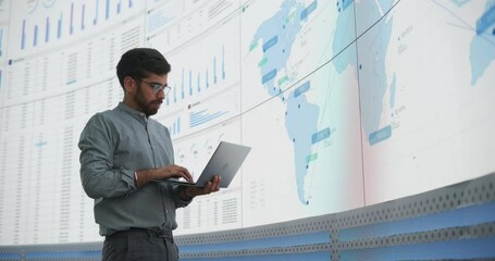 Wall Mural - Indian Male Logistics Expert Holding Laptop Computer And Analyzing World Map On Big Digital Screen In Monitoring Office. Successful Man Developing New Efficient Routs For Global Product Distribution.