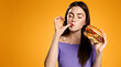 Brunette girl licks her finger, eats tasty hamburger. Woman orders takeaway food with delivery app, enjoys delicious burger, orange background