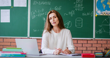 Attractive female math teacher sitting at desk with laptop in modern classroom. Confident woman talking and holding paper sheet with triangle while looking at camera.