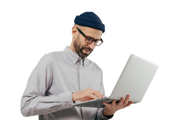 Concentrated bearded male administrative worker coordinates work on distance, searches information on laptop computer, wears white formal shirt and transparent glasses, poses indoor over grey wall