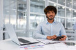 © Liubomir - Portrait of young successful financier accountant at work inside office building at workplace, hispanic man smiling and looking at camera, man using calculator in paperwork.
