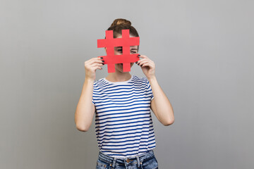 Wall Mural - Portrait of unknown woman wearing striped T-shirt looking through red hashtag sign, looking for proper posts in social media, spying. Indoor studio shot isolated on gray background.