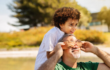 Wall Mural - Cheerful old caucasian grandfather holds small mixed race boy on his shoulders, closes eyes to man