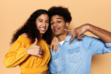 Studio image of two happy people, boyfriend and girlfriend, gesticulating in front of camera against brown background, girl showing thumb up and boy making victory sign, feeling successful and cool