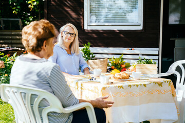 Cheerful aged senior woman drinking tea, talking to her adult daughter in rural country house garden outdoor on sunny autumn day.