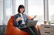 © sofiko14 - Young pretty female in jeans and jacket having video conference on laptop and spreading hands in glassy office. Attractive short haired woman in spectacles sitting on chair and talking to coworkers.