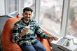Portrait of nice-looking indian employee sitting on soft office chair with personal laptop on knees and smiling at camera. Imposing dark haired man taking short break after intensive working day.