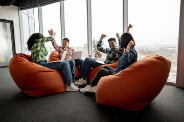 Diverse team of excited professionals in street style clothes raising fists in air and giving high five in workspace interior. Happy managers with devices celebrating successful project launch.