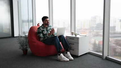 Handsome indian man in casual attire speaking on video call on laptop and gesturing hands in cozy office. Young bearded employee sitting in white spacious workplace with great panoramic view on city.