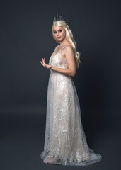 Wall Mural - Full length portrait of beautiful women with long blonde hair, wearing fantasy  princess crown and elegant white ball gown, standing pose with hand gesture. Isolated on dark grey studio background.