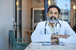 © Cavan Images - Portrait of male doctor wearing eyeglasses sitting at desk in clinic