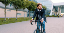 Likable Thoughtful Young Bearded Businessman With Backpack In Formal Clothes Riding A Bicycle To His Office On Cobbled Street Near Big Panoramic Building