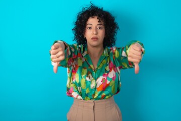 young arab woman wearing colorful shirt over blue background being upset showing thumb down with two hands. Dislike concept.