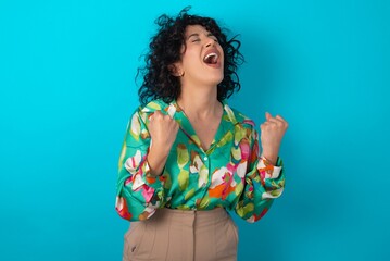 Sticker - young arab woman wearing colorful shirt over blue background celebrating surprised and amazed for success with arms raised and eyes closed. Winner concept.