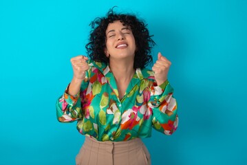 Wall Mural - young arab woman wearing colorful shirt over blue background being excited for success with raised arms and closed eyes celebrating victory. Winner concept.