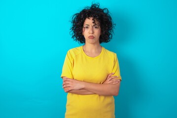 Gloomy dissatisfied young arab woman wearing yellow T-shirt over blue background looks with miserable expression at camera from under forehead, makes unhappy grimace