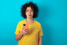 Upset Dissatisfied Young Arab Woman Wearing Yellow T-shirt Over Blue Background Uses Mobile Software Application And Surfs Information In Internet, Holds Modern Mobile Hand
