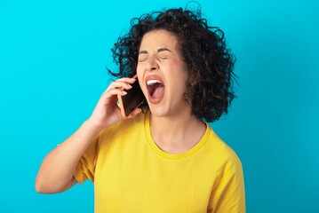 Sticker - Overemotive happy young arab woman wearing yellow T-shirt over blue background laughs out positively hears funny story from friend during telephone conversation