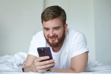 Canvas Print - Young bearded happy man is using cell mobile phone at home in bed in bedroom, looking at screen of smartphone 