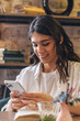 © Tatyana - Portrait of a smiling dark-haired young woman using a smartphone in a cafe.Remote work,business, freelance,blogging,social media concept.
