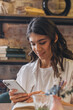 © Tatyana - Portrait of a smiling dark-haired young woman using a smartphone in a cafe.Remote work,business, freelance,blogging,social media concept.
